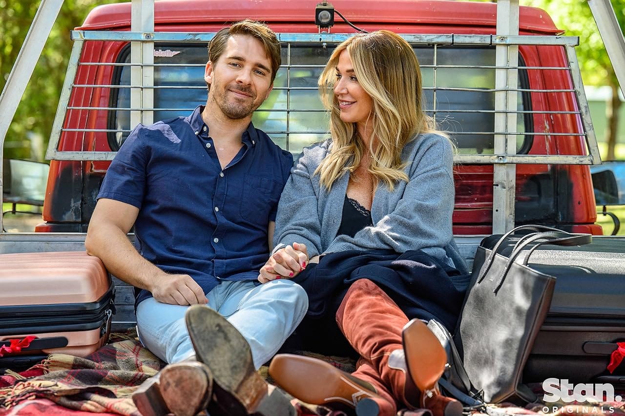 Man in blue collared shirt, jeans and boots sitting in back of ute with women in cardigan, skirt and boots