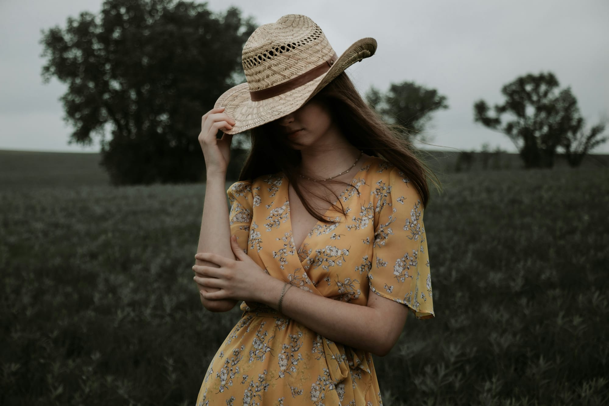 Women wearing yellow floral dress with hat pulled over face, countryside in the background