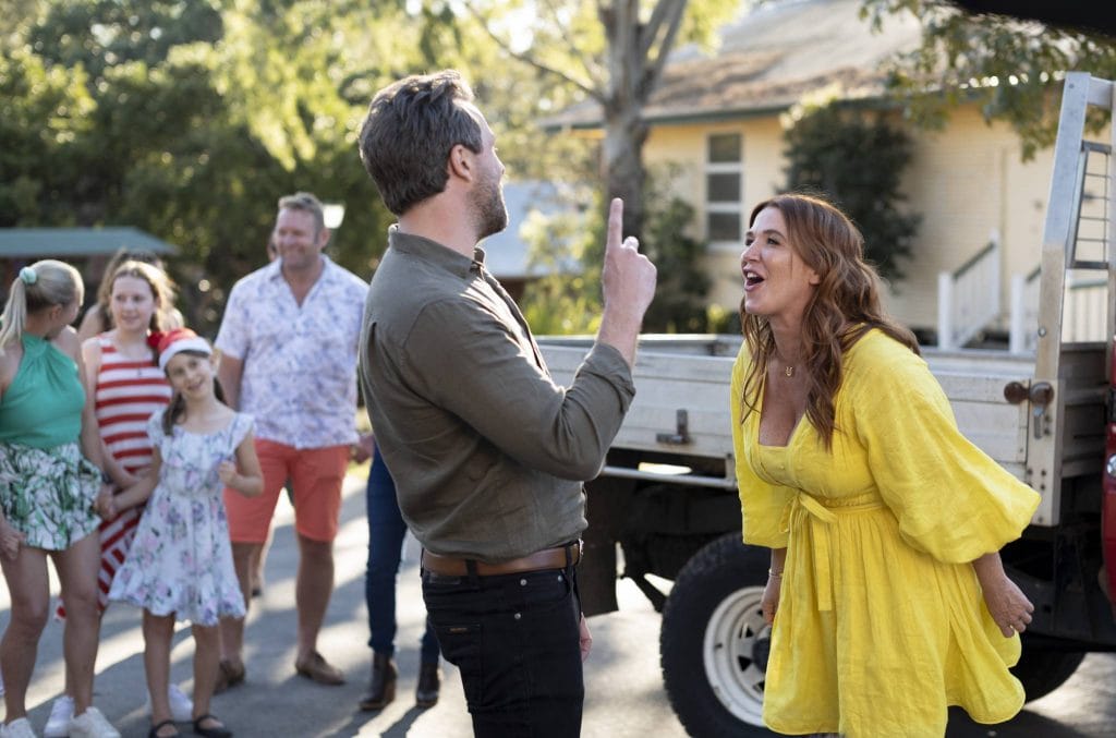 Women in yellow dress next to car dancing and singing with man with family looking on